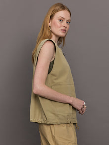 Model wears a wide shoulder, khaki zip up vest with tactical symmetrical front cargo pockets at the waist.