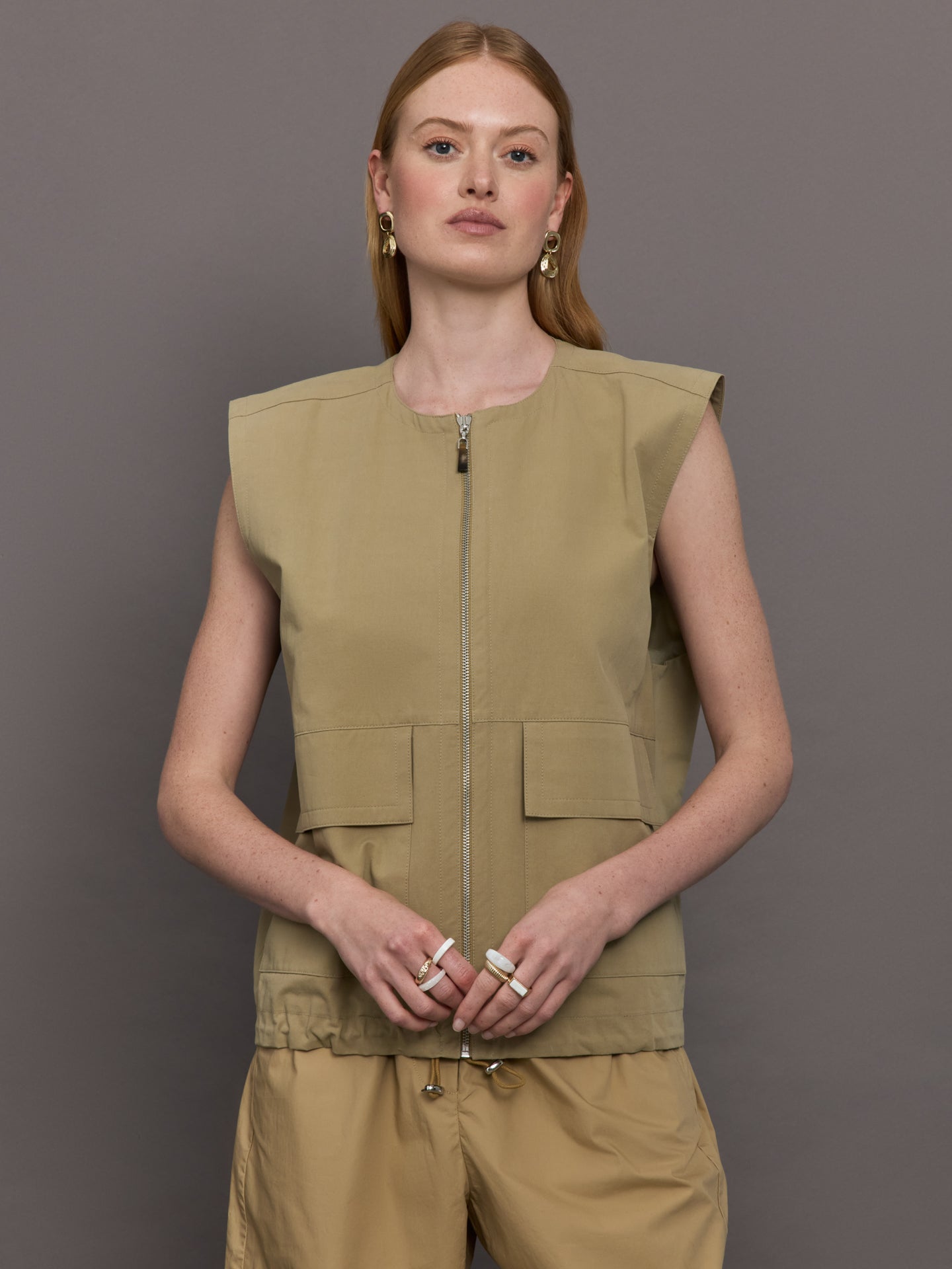 Model wears a wide shoulder, khaki zip up vest with tactical symmetrical front cargo pockets at the waist.