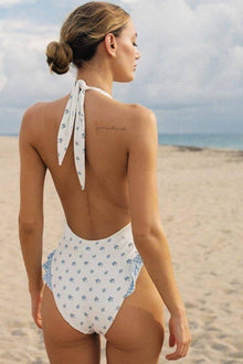 Woman wearing white one-piece swimsuit with blue floral print and halter neckline, shown from back on beach.