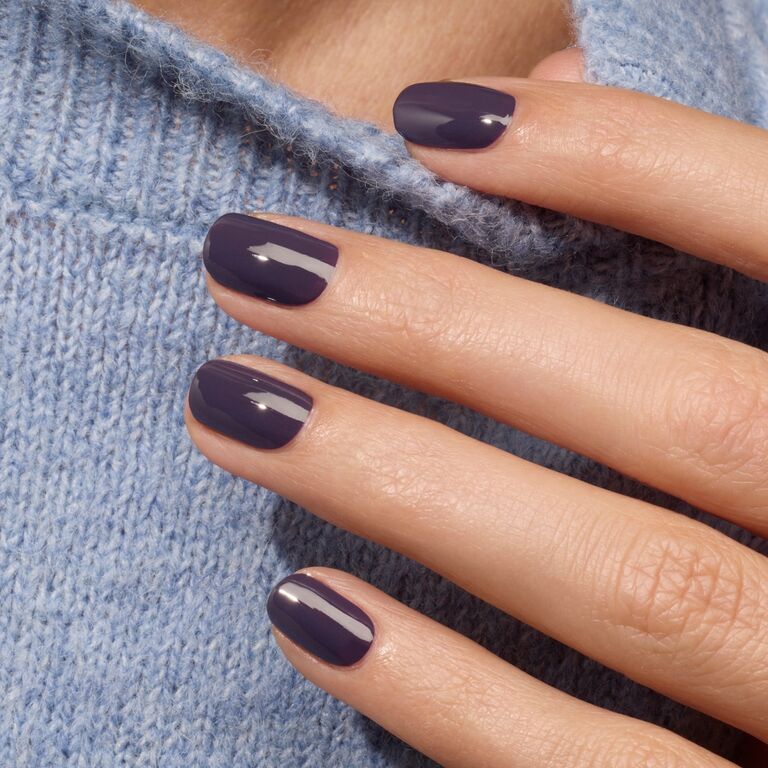 A close-up of a hand with neatly manicured nails painted in Manucurist's Queen of Night deep purple polish, resting on a light blue knitted sweater.