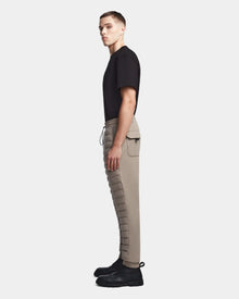 A man stands in profile against a light background, wearing a black T-shirt, Rudsak MIKE - 6523503 Cement mens quilted taupe pants with pockets, and black lace-up boots.
