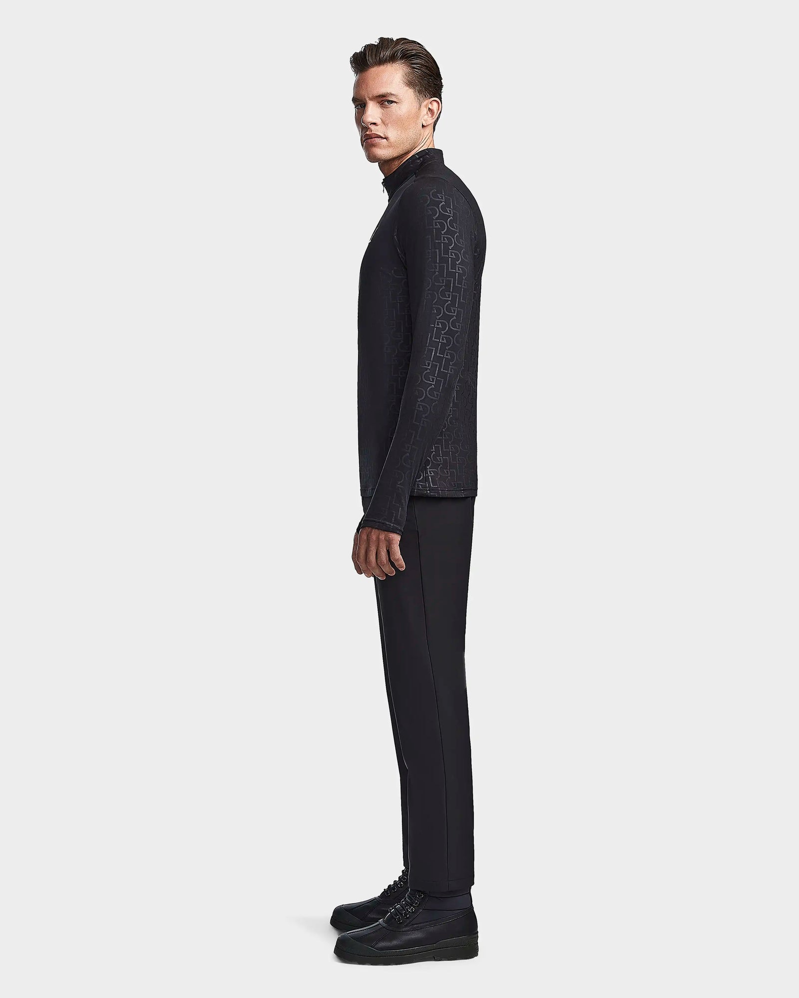 A man stands in profile against a plain background, wearing the Rudsak WARREN - 6523502 Black Pattern mens fleece-lined fitted stretch top, paired with black pants and shoes.