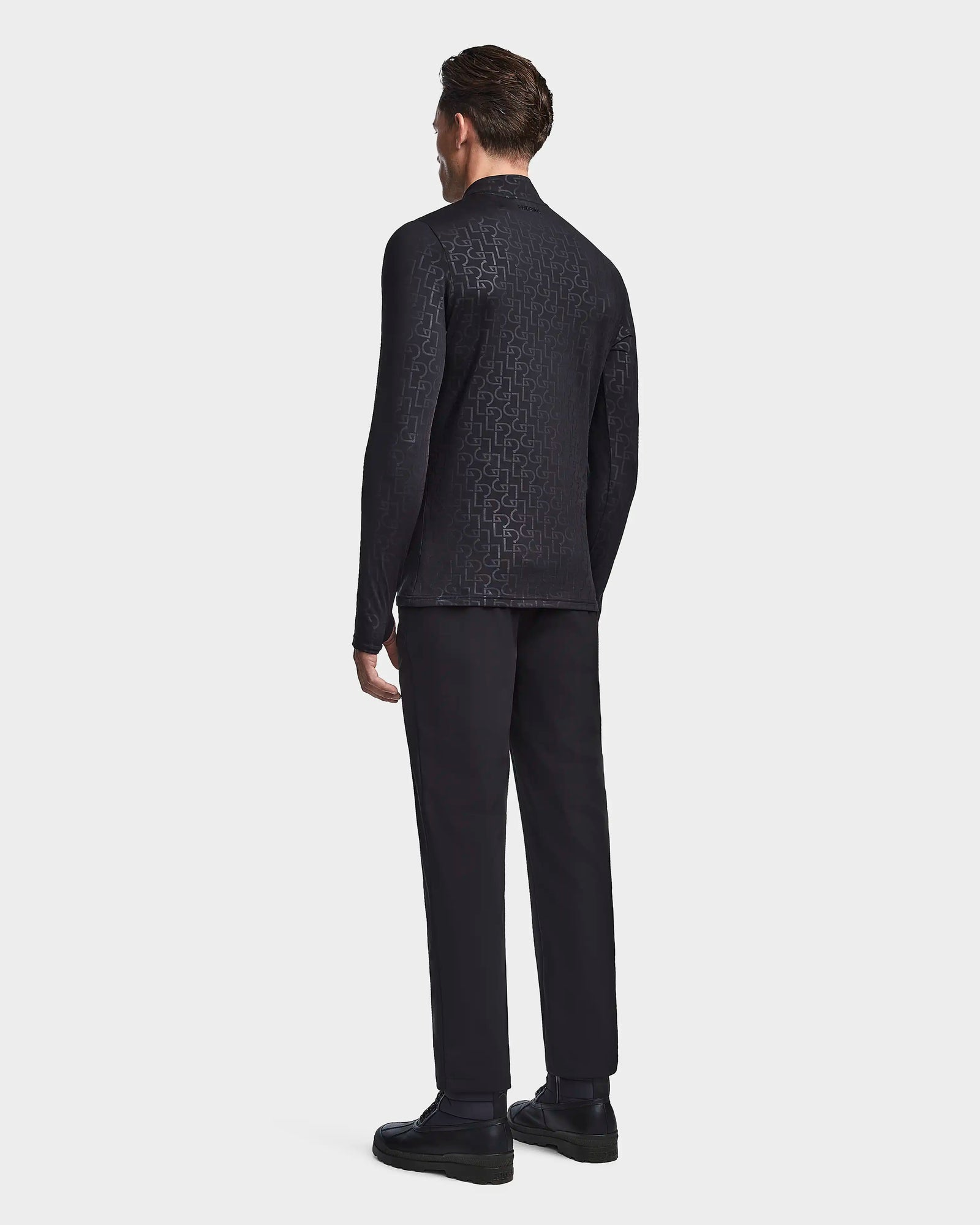 A man faces away from the camera, wearing the Rudsak WARREN - 6523502 Black Pattern fleece-lined zip-front mock neck jacket, black pants, and black shoes against a plain light gray background.