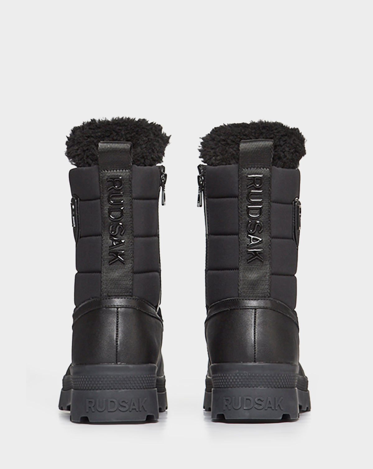 The REAGAN - 6222500 BLACK men’s winter boots by Rudsak feature black leather, thick soles, quilted uppers, and black faux fur lining. Rudsak branding appears on the heel, pull tab, and side. Shown from the back.