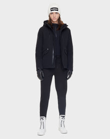 A person stands facing forward against a plain light gray background, wearing the Rudsak DEST - CA EN 6119516 BLACK goose down winter parka, white beanie, black gloves and pants, and white lace-up boots.