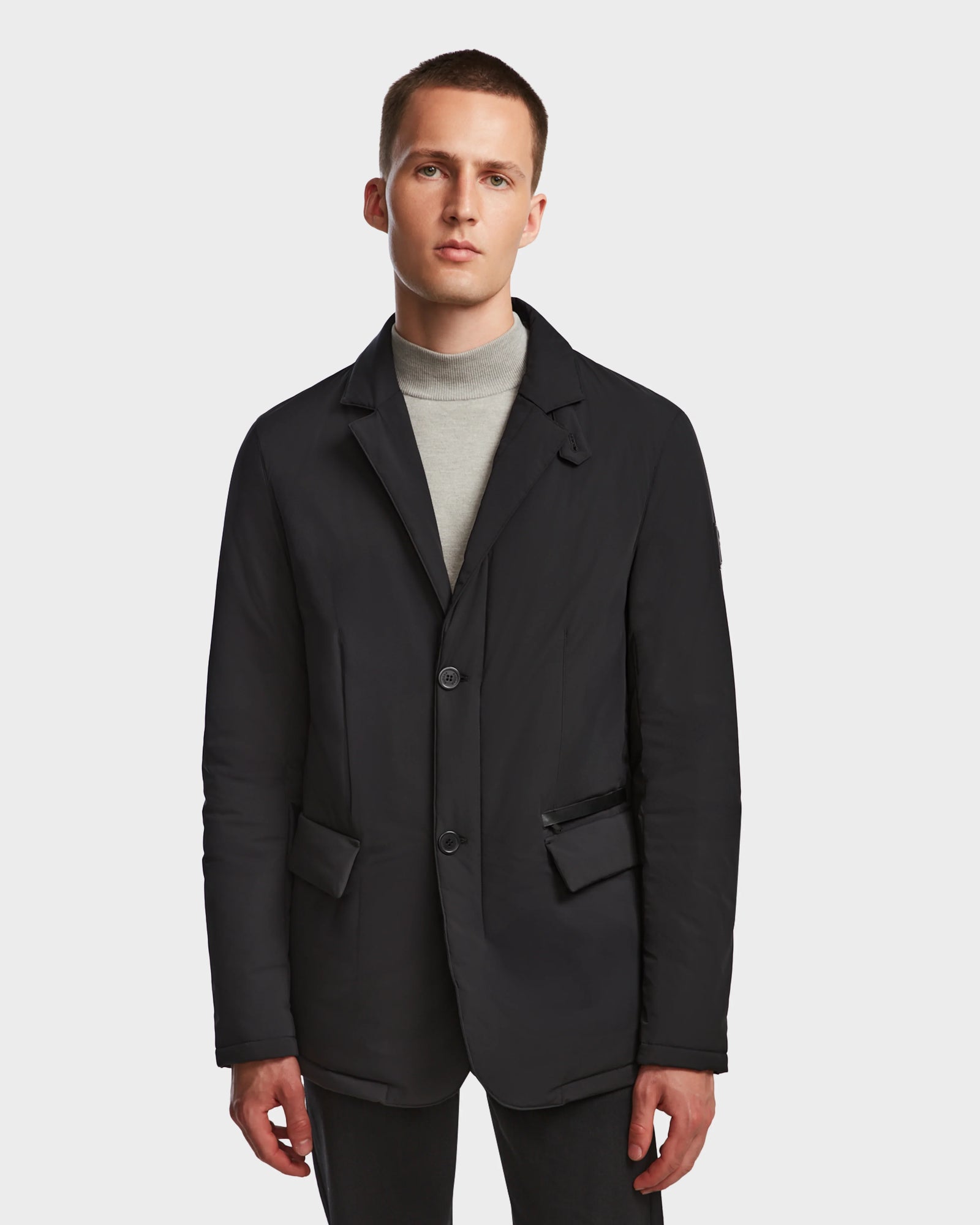 A young man with short hair wears the Rudsak OTTO - 6124602 Black mens blazer-style down jacket with eco polyfill over a light gray turtleneck, standing against a plain white background.