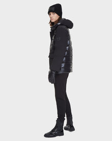A person stands slightly to the side against a plain white background, wearing the Rudsak DUSK - M121506 BLACK men's puffer jacket with waterproof zippers, paired with a black beanie, gloves, fitted pants, and lace-up boots.