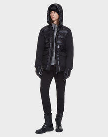 A person wearing the Rudsak DUSK - M121506 BLACK men's puffer jacket with waterproof zippers, paired with black pants, gloves, boots, and a checkered shirt, stands against a plain white background.