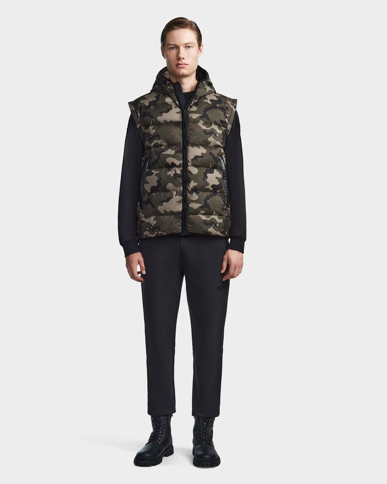 A person stands facing forward, wearing the Rudsak DEMIAN M-R - M123504 Camouflage mens down puffer vest over a black long-sleeve shirt, black pants, and lace-up boots against a plain light background.
