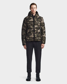 A person stands facing forward wearing the Rudsak DEMIAN M-R - M123504 Camouflage mens down puffer, paired with black pants and black lace-up boots against a plain light background.