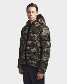 A person in Rudsaks DEMIAN M-R - M123504 Camouflage mens down puffer jacket with a hood and black zipper details, paired with black pants, stands against a plain white background.