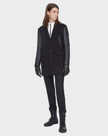 A person with long brown hair wears the Rudsak BEAU - M121504 BLACK men’s coat, styled with black pants, gloves, and boots. They stand slightly angled against a plain white background.