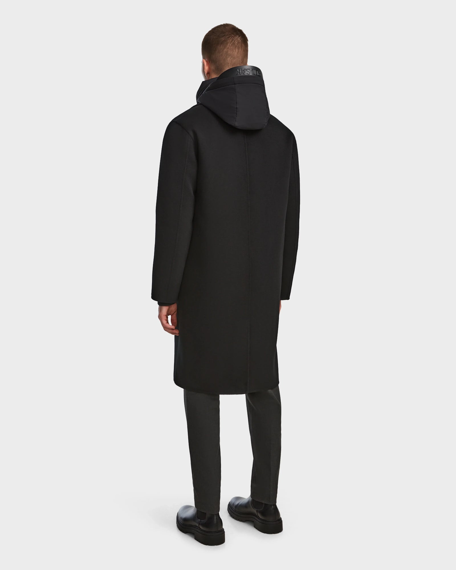 A man faces away, wearing the Rudsak DRAL - 6124503 Black wool jacket with a hood, paired with dark pants and black shoes, set against a plain light background.