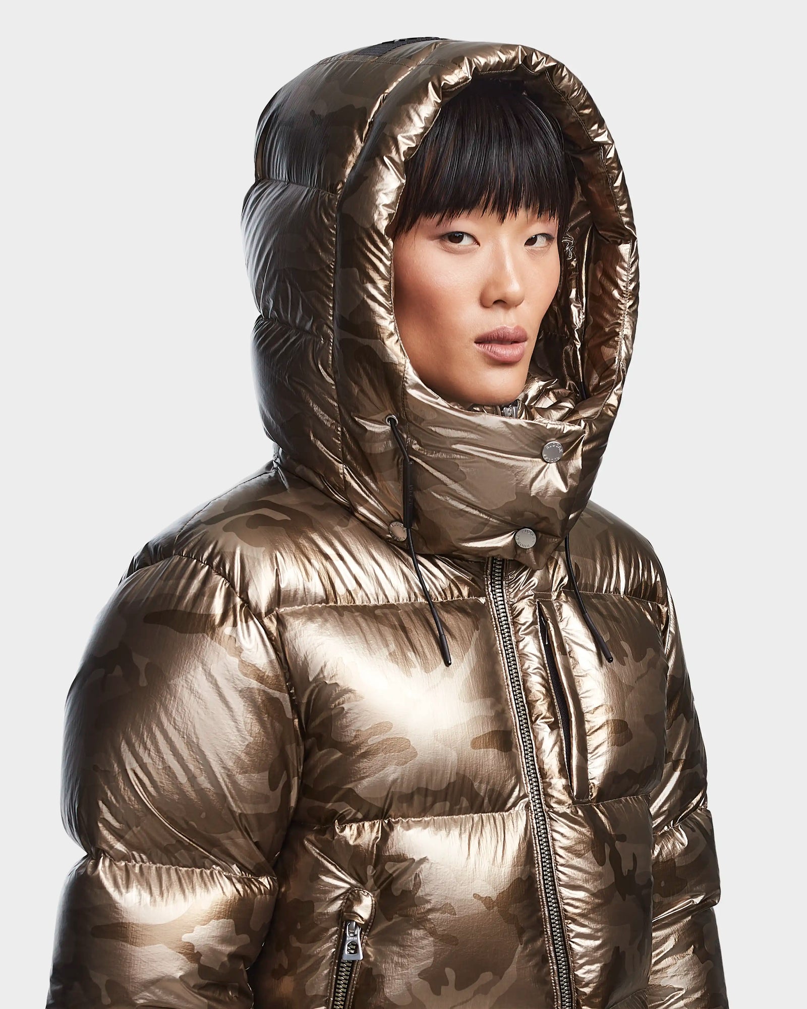 A person wearing the Rudsak MAVE S - 8123563 Metallic Ivory Camo womens down puffer jacket with a large hood up, looking at the camera against a plain light background.