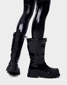 A person wears Rudsaks KRISTAL HI - 8222510 BLACK womens boots, featuring high-gloss finish, chunky grip soles, and RUDSAK branding at the back, standing against a plain light background.