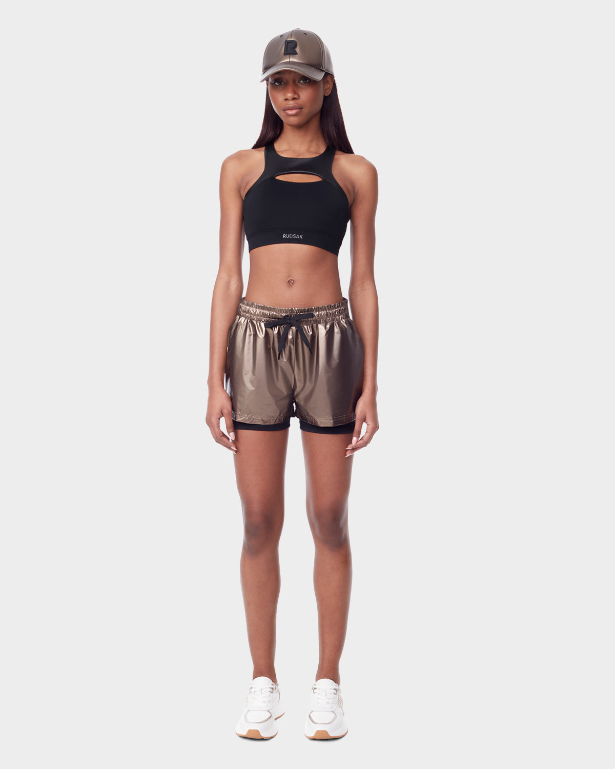 A woman wears Rudsaks RYLEE - 8523001 METALLIC CHOCOLAT glossy bronze workout shorts with a black crop top, white sneakers, and a matching bronze cap, posing against a plain light background.