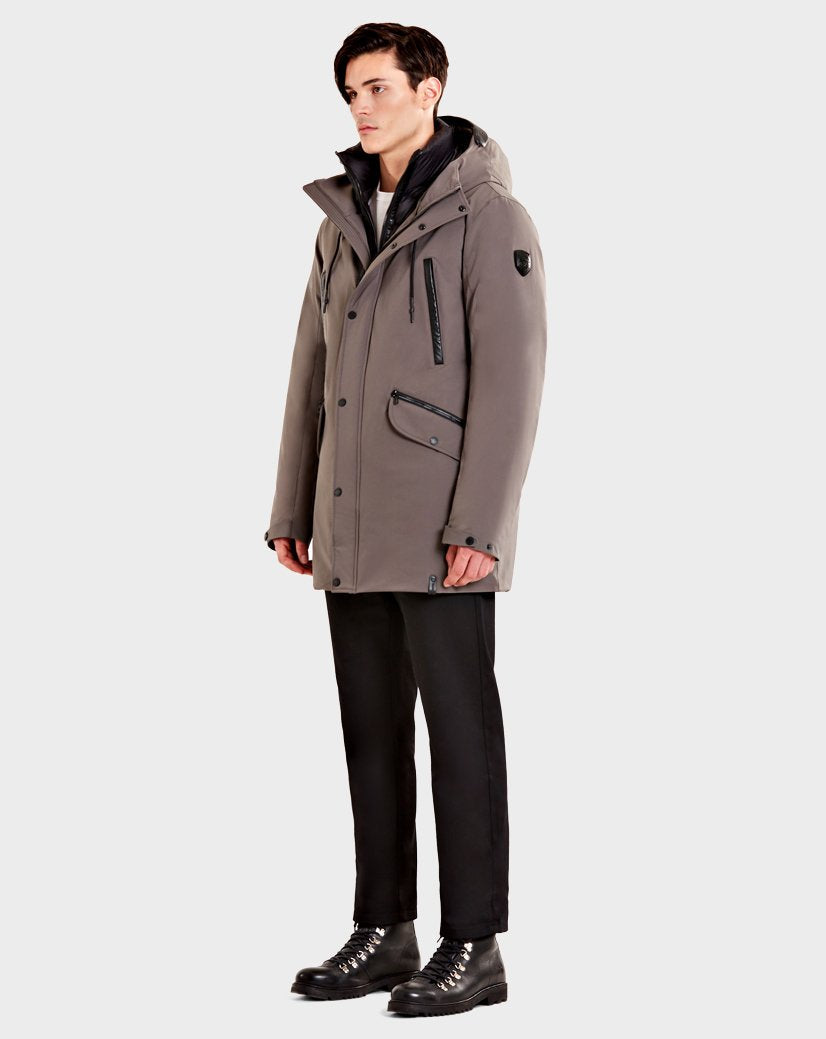 A man stands facing left, wearing the Rudsak DENT - CA EN 6119515 ELEFANTE gray mid-length parka, black pants, and black lace-up boots against a plain light gray background.