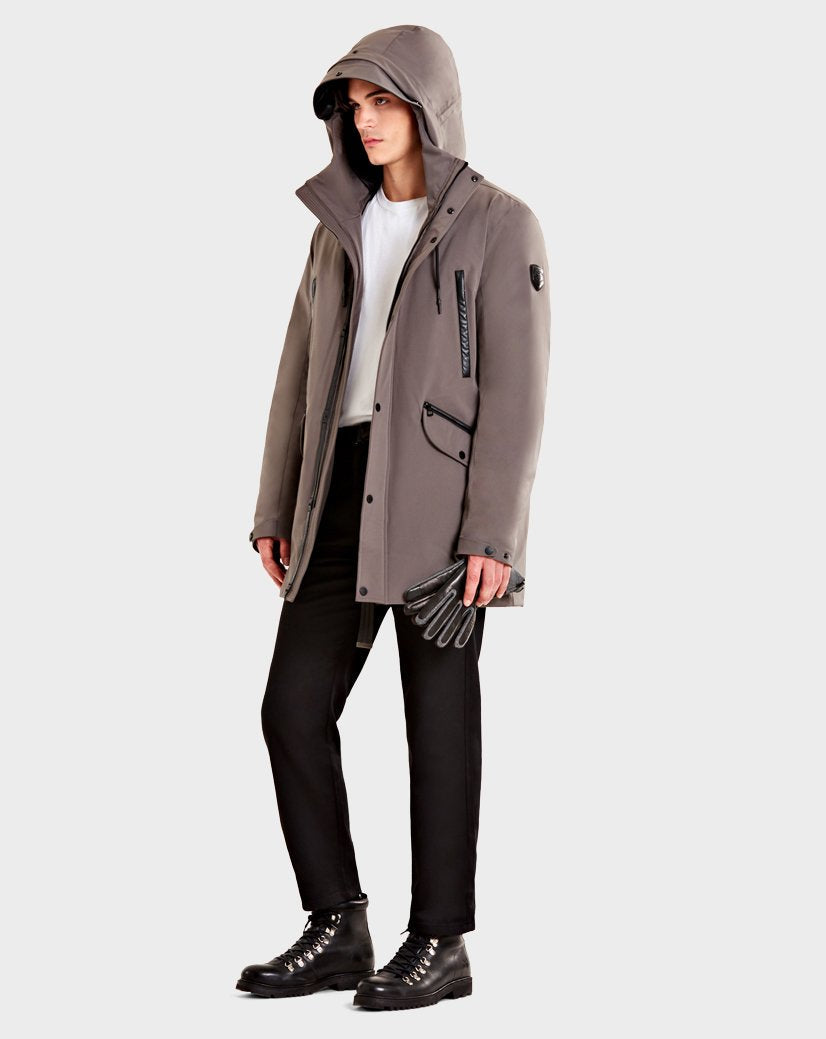 A person wearing the Rudsak DENT - CA EN 6119515 ELEFANTE gray mid-length parka with leather trim, white shirt, black pants, and black boots stands against a plain light background, holding black gloves in one hand.