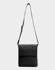 The Rudsak MADDY - 8322512 BLACK is a sleek black leather messenger bag with a long adjustable strap and front flap closure, displayed in minimalist style against a plain light gray background.