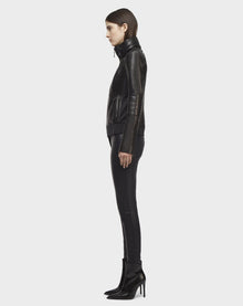A person with straight brown hair models the Rudsak PRESLEY - CA EN 8119029 BLACK lamb leather biker jacket, black skinny pants, and black heeled boots, standing in profile facing left against a plain white background.