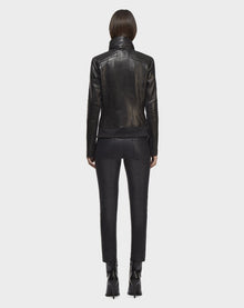 A person with straight, shoulder-length hair faces away, wearing the Rudsak PRESLEY - CA EN 8119029 BLACK biker jacket, paired with black pants and heeled boots, standing against a plain light background.