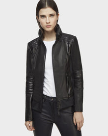 A woman with straight brown hair wears the Rudsak PRESLEY - CA EN 8119029 BLACK lamb leather biker jacket over a white shirt and black pants, standing against a plain light gray background.