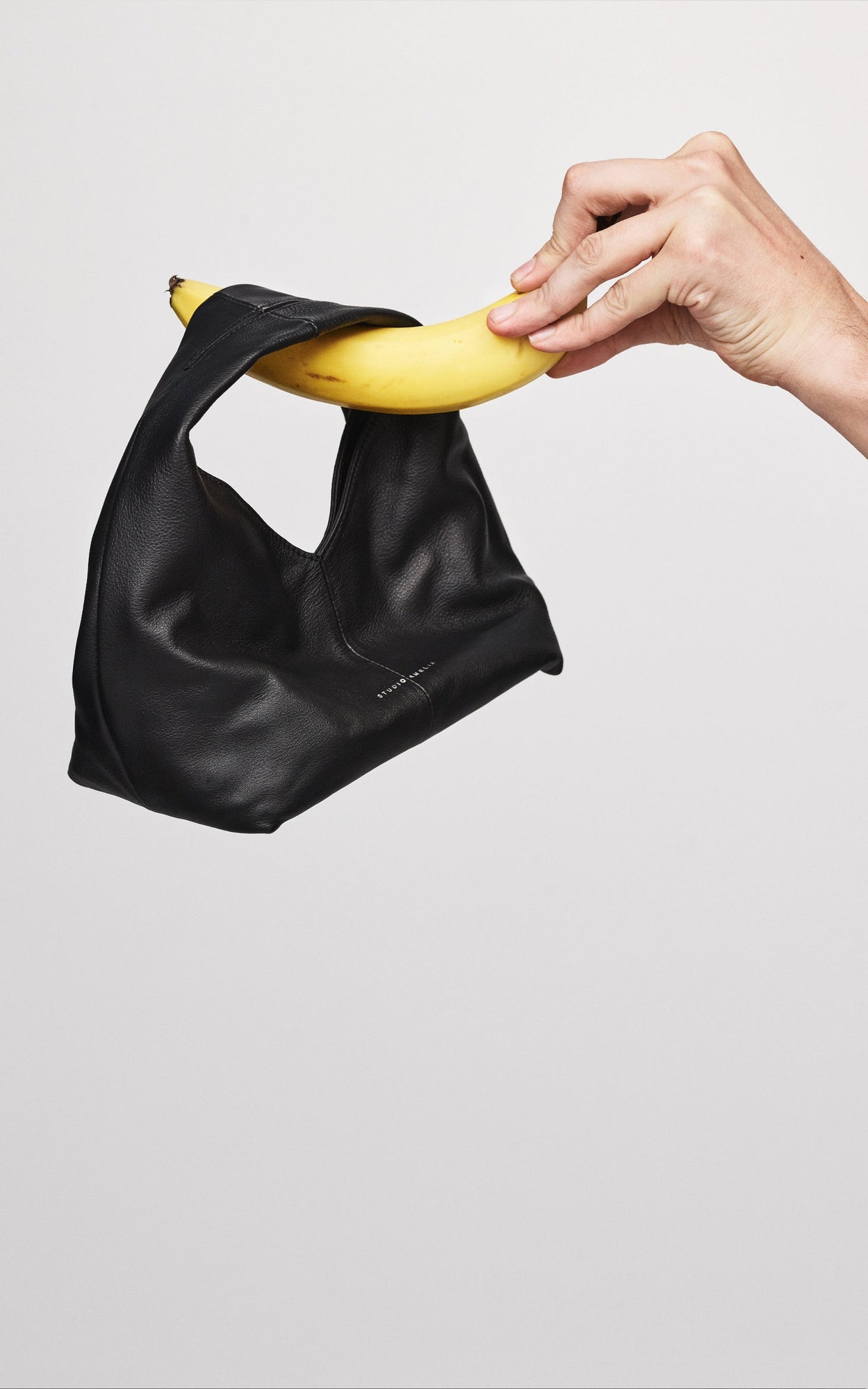 Black handbag with a banana being held by a hand against a light gray background