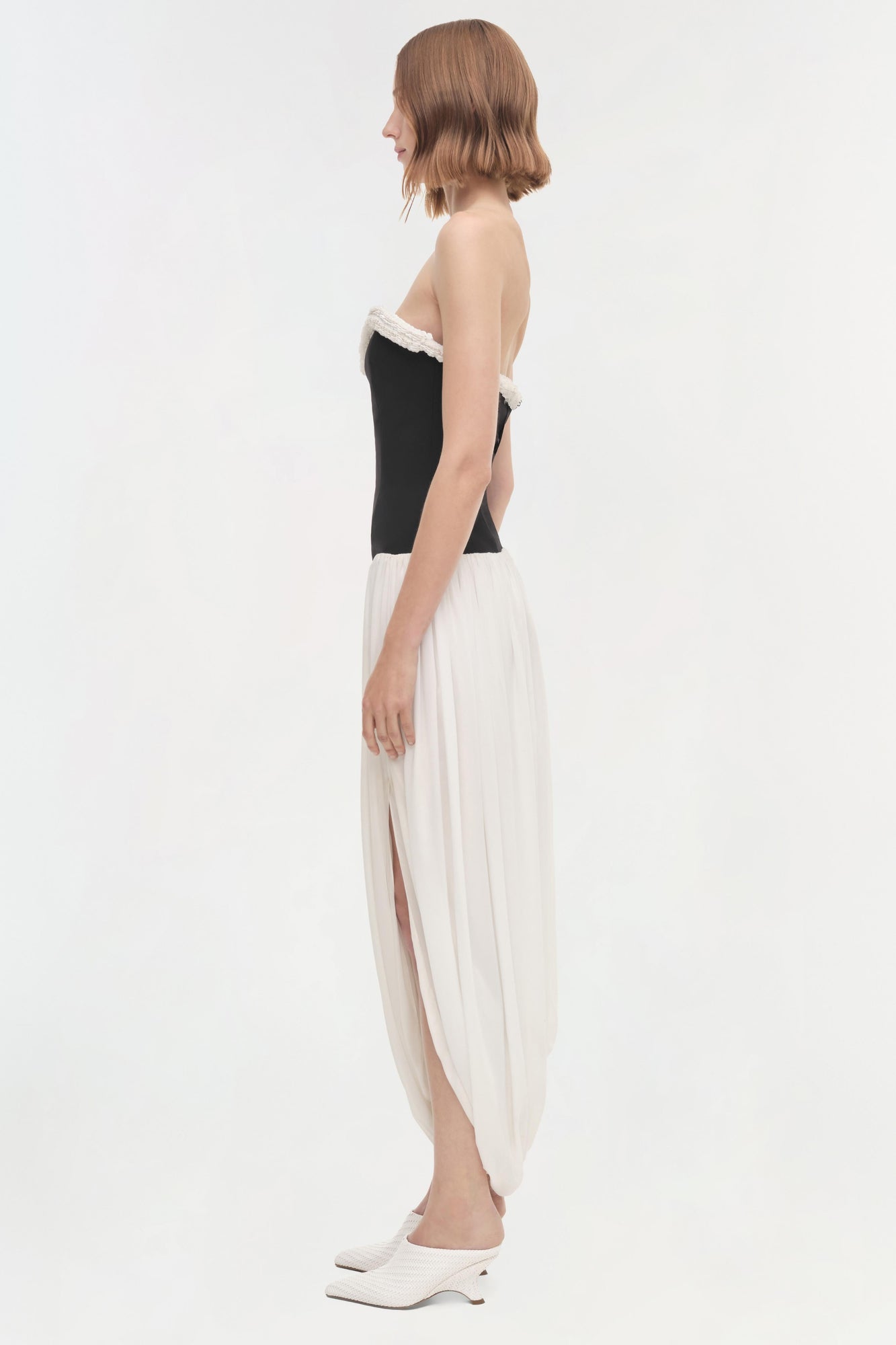 Woman wearing strapless dress with black bodice, white lace trim, and white pleated skirt, shown from side profile.