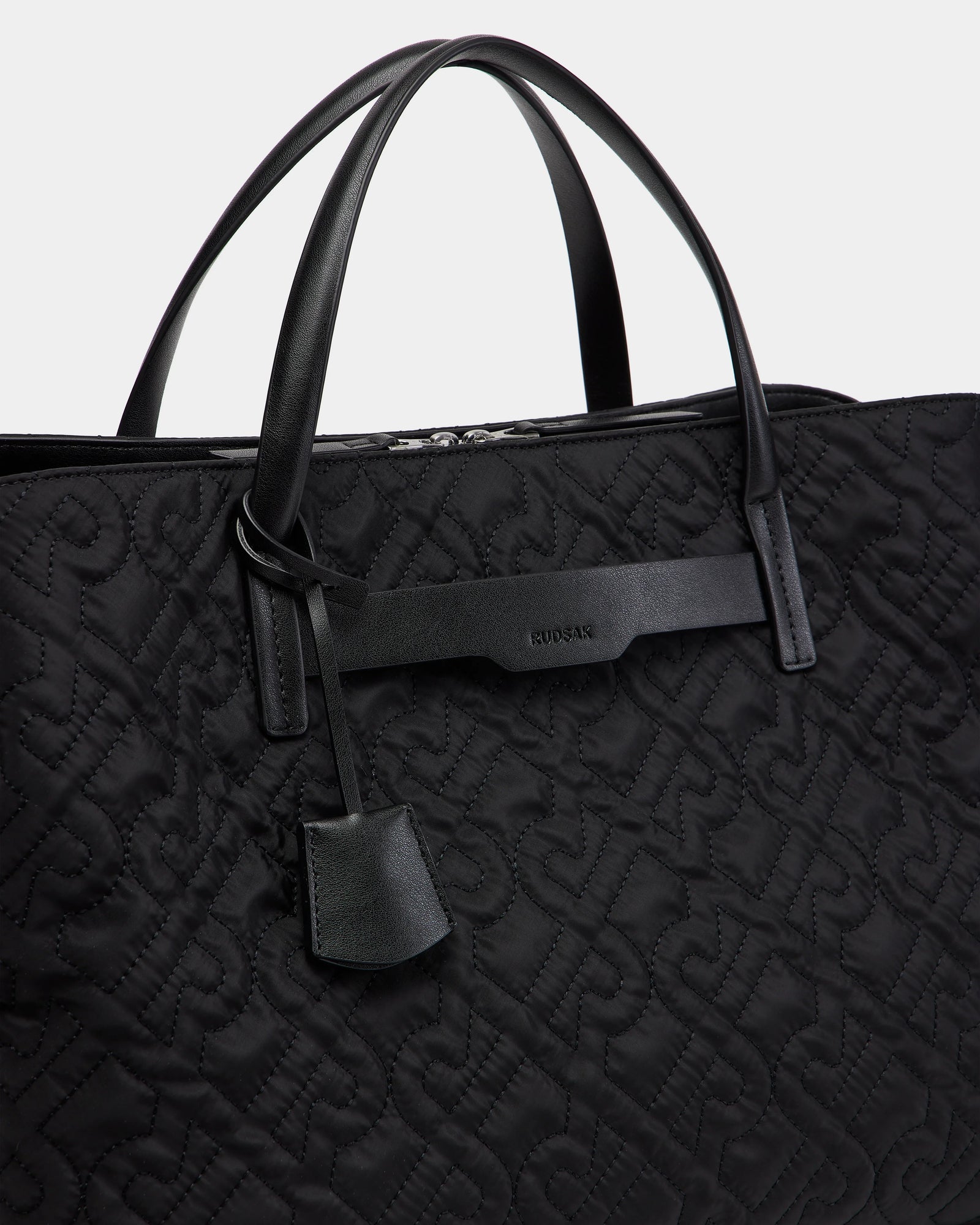 Close-up of the Rudsak ASMINE - L325005 Black tote with an embossed monogram pattern, two leather handles, a leather tag, and a branded leather accent near the top.