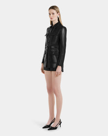 A woman with long blonde hair and a neutral expression wears the Rudsak DANICA - 8125025 Black womens leather jacket, styled with front pockets, black shorts, and black high heels against a plain white background.