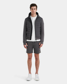 A man faces forward against a plain white background, wearing the Rudsak DEE MAT - 6124028 Grey mens lightweight down jacket, paired with a white t-shirt, gray shorts, and white sneakers.