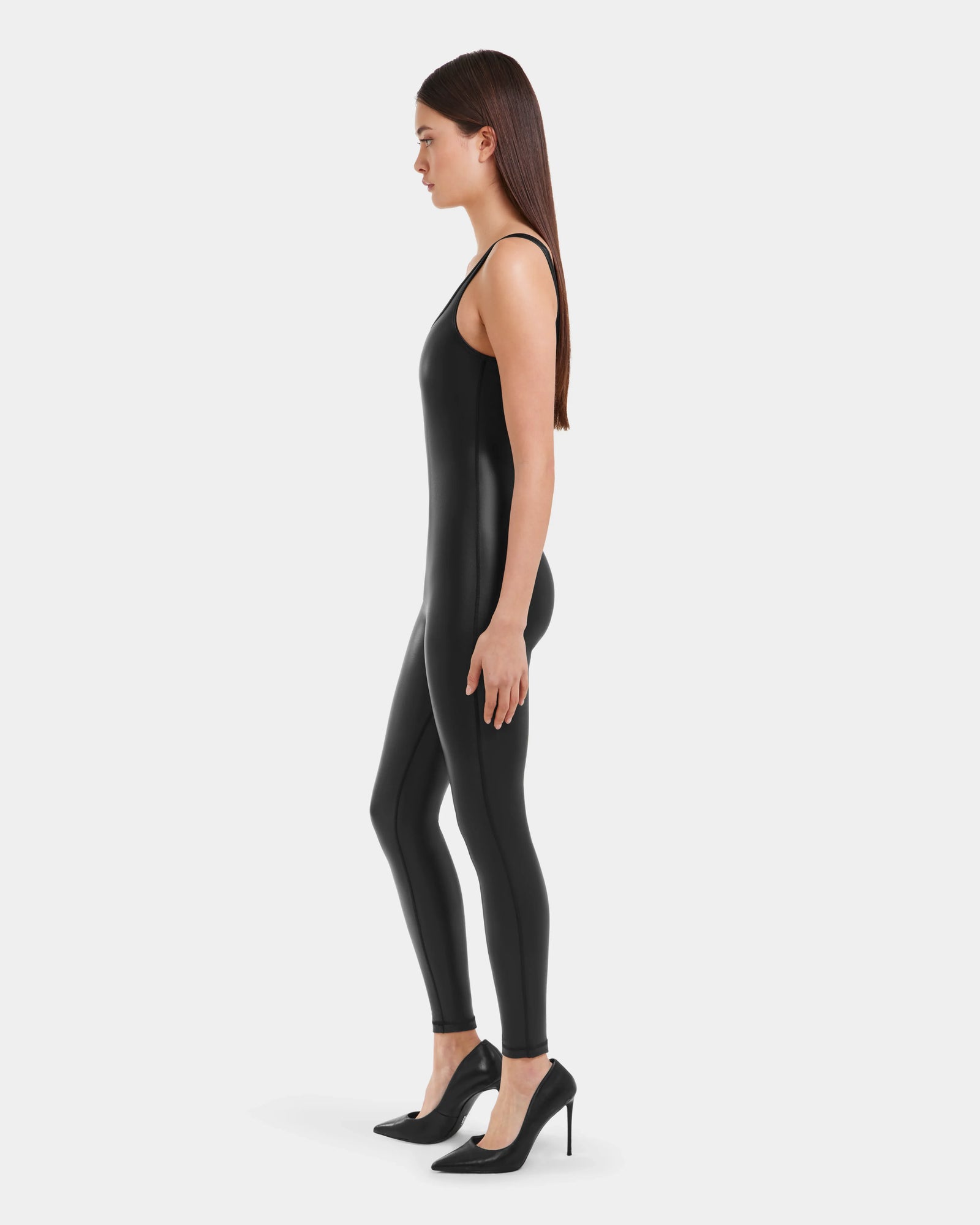 A woman with long brown hair stands in profile against a light gray background, wearing the Rudsak LAWREN - 8523047 Black sleeveless bodysuit and black high-heeled shoes.