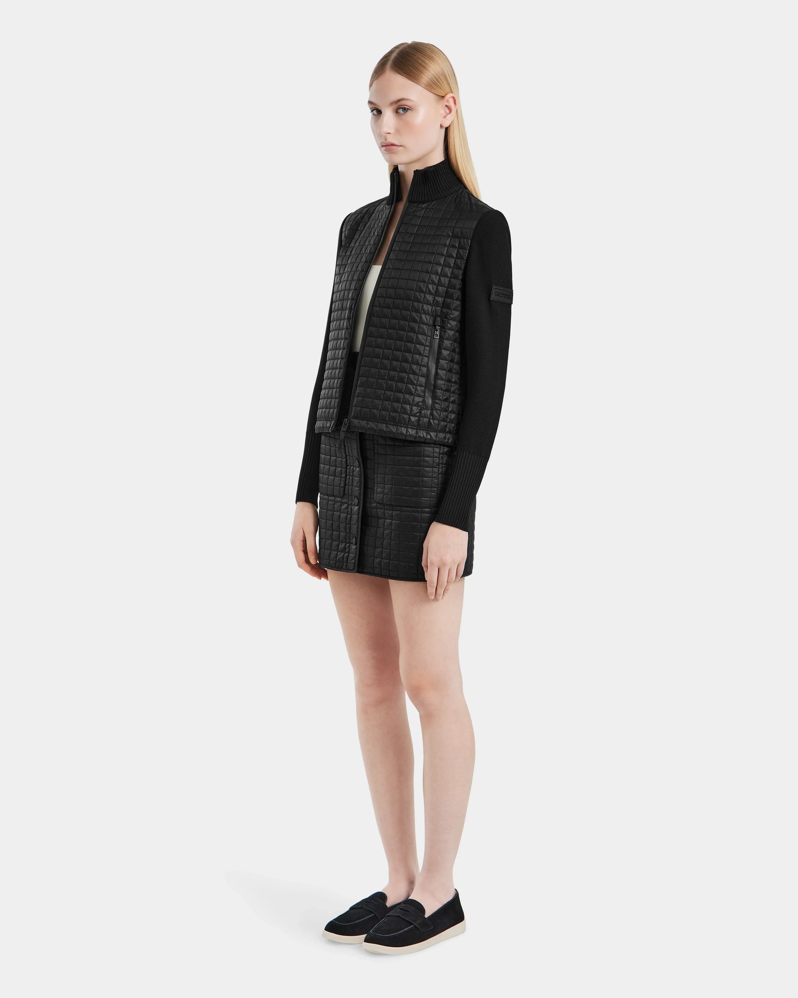 A woman stands slightly to the side, wearing Rudsaks LOUISE - 8124046 Black womens hybrid knit sweater jacket and matching mini skirt with black slip-on shoes against a plain white background.