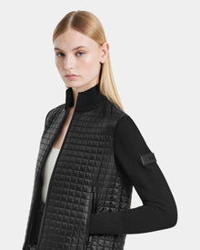 A woman with straight blonde hair models the Rudsak LOUISE - 8124046 Black womens hybrid knit sweater jacket, featuring RDS-certified down, lightweight quilting, knit sleeves, and a standing collar. She poses sideways against a plain light background.