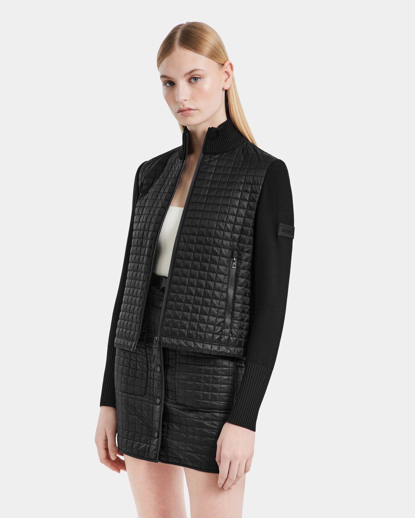 A woman with straight blonde hair wears the Rudsak LOUISE - 8124046 Black lightweight quilted jacket and matching skirt over a white top, standing against a plain light gray background.