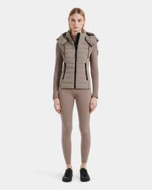 A woman faces forward, wearing Rudsaks MABS - 8124825 Cement taupe puffer vest over a lightweight jacket, paired with a long-sleeve taupe top, matching fitted pants, and black sneakers against a white background.