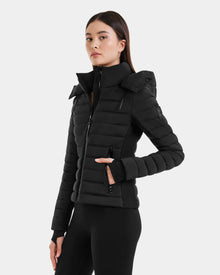 A woman with straight dark hair wears the Rudsak MARYA M - L123559 Black women’s down puffer jacket, featuring a sleek design, high collar, and zippered pockets, while standing against a plain light background.