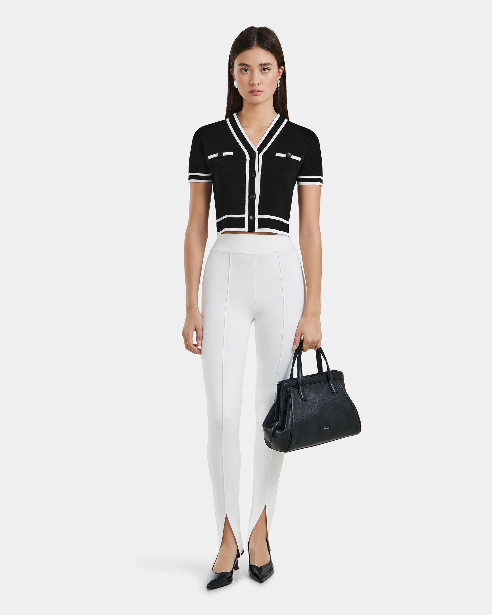 A woman faces forward wearing a black short-sleeve buttoned top, Rudsak’s MATHILDA - 8524515 Iceland front ankle slit leggings, black pointed-toe heels, and holding a black handbag.