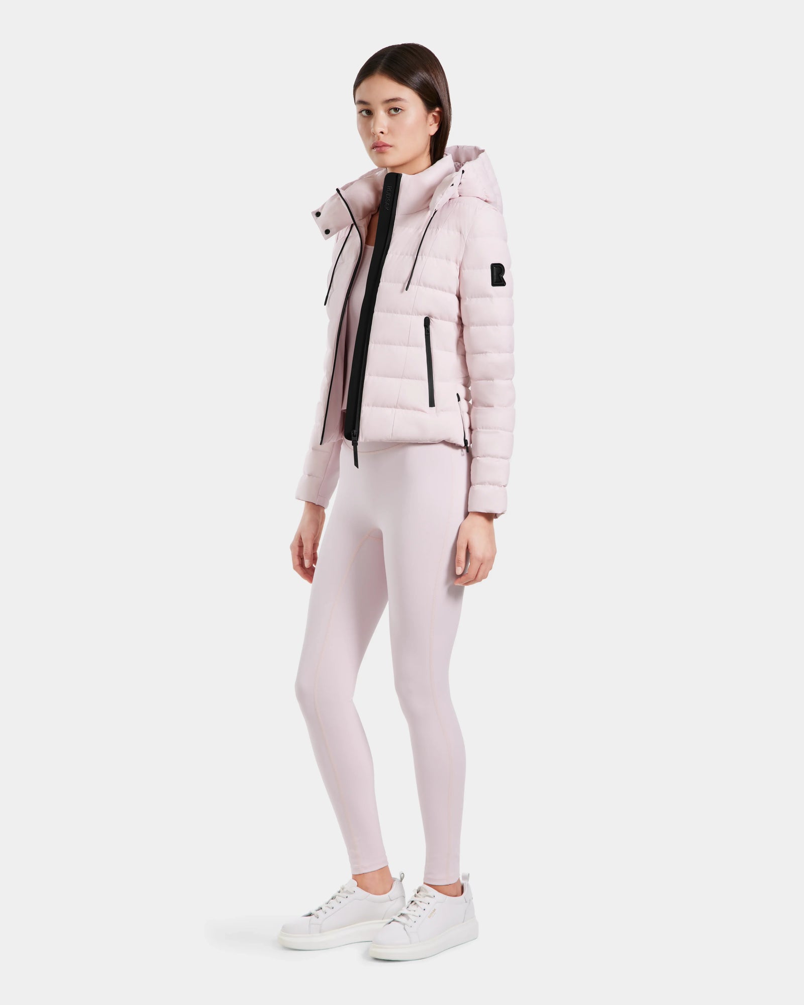 A woman with straight dark hair and a neutral expression poses against a plain background, wearing the Rudsak MELISSA - 8125054 Light Pink puffer jacket, matching leggings, a white top, and white sneakers.