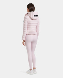 A woman faces away, wearing the Rudsak MELISSA - 8125054 light pink puffer jacket with a hood, light pink leggings, and white sneakers against a plain white background.