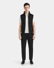 A man faces forward against a light gray background, wearing the Rudsak MERLO - 6124017 Black quilted vest over a white t-shirt, paired with black pants and black sneakers with white soles.