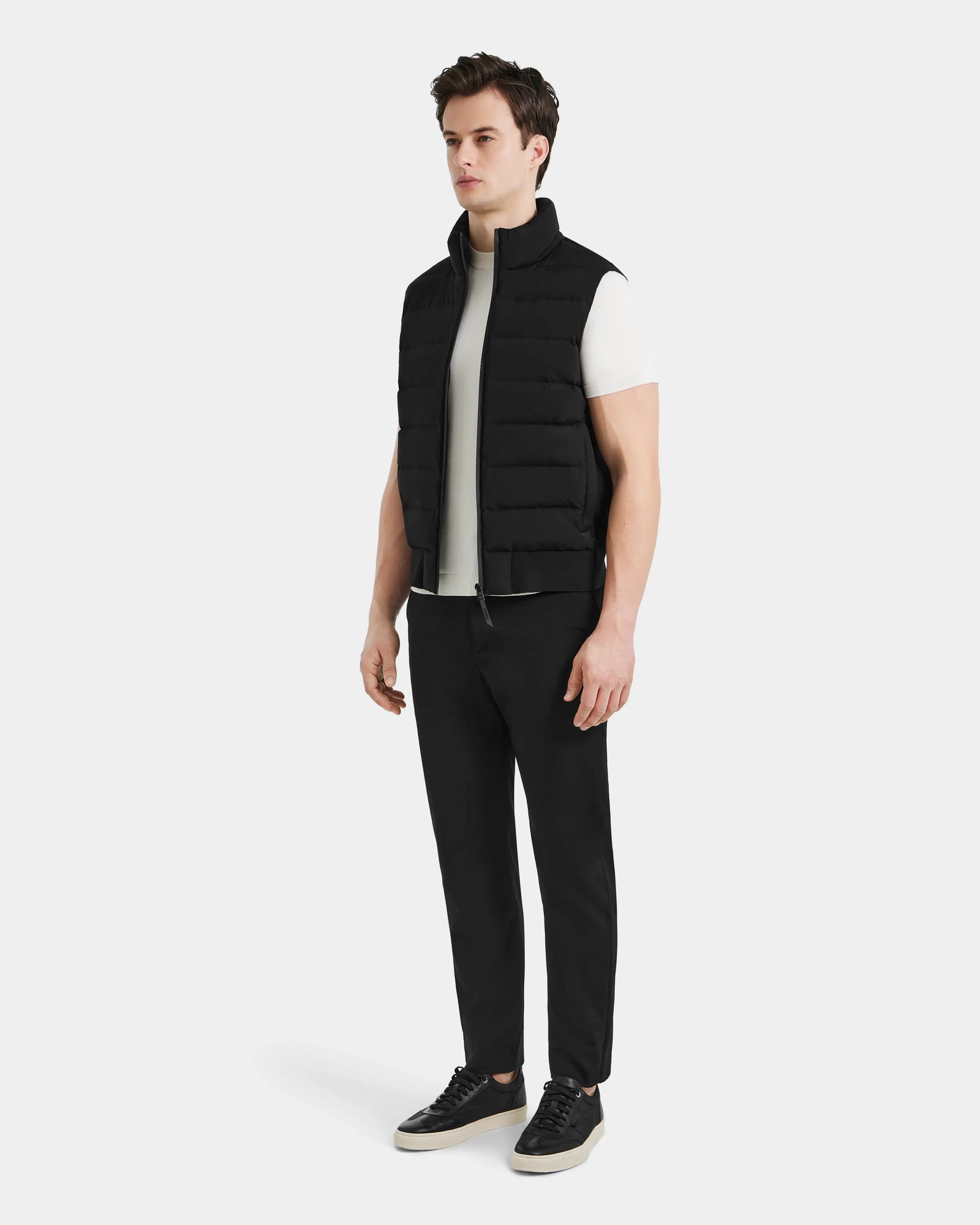A man stands against a plain background wearing a white T-shirt, black pants, black sneakers, and the Rudsak MERLO - 6124017 Black mens quilted vest. He faces forward with a neutral expression.