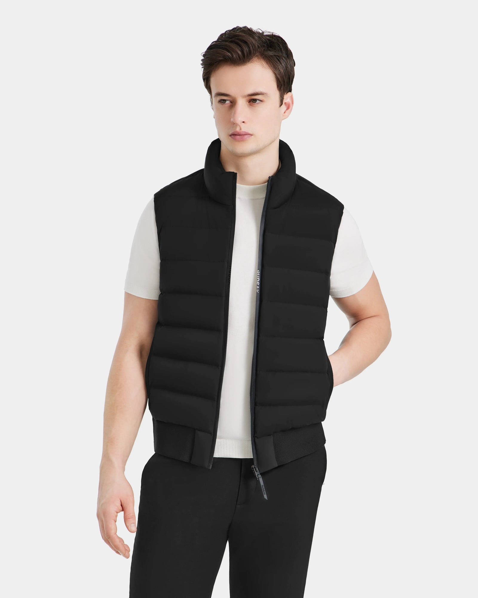 A young man stands against a plain light background, wearing the Rudsak MERLO - 6124017 Black quilted vest over a white t-shirt and black pants, one hand in his pocket as he looks slightly to the side.