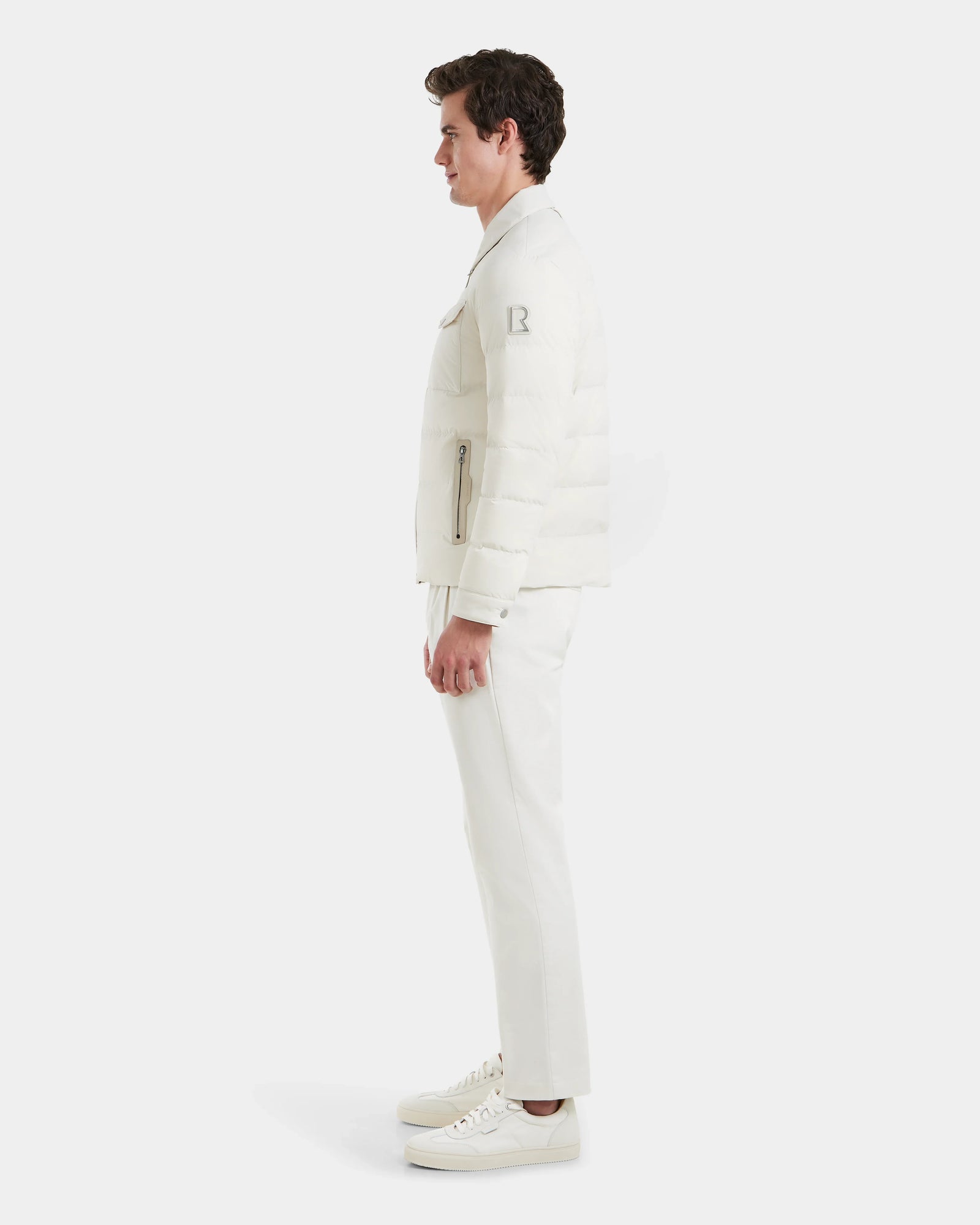 A man stands in profile against a white background, wearing the Rudsak ORNO - 6124601 Vanilla mens down quilted jacket with RDS-certified down, paired with white pants and sneakers.