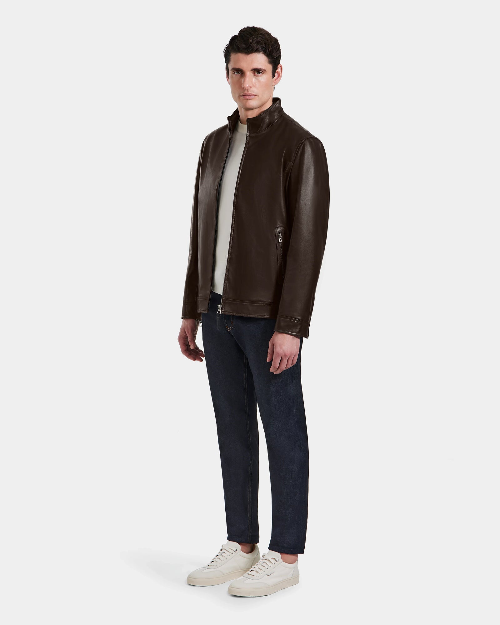 A man faces forward, wearing the Rudsak STEVEN - 6124541 Espresso mens leather jacket with a white shirt, dark blue jeans, and white sneakers, set against a plain light gray background.