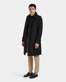 A man faces forward with a neutral expression, wearing the Rudsak TIMOTHY - 6124589 Black knee-length trench coat over a black shirt, tan pants, and black loafers against a light background.