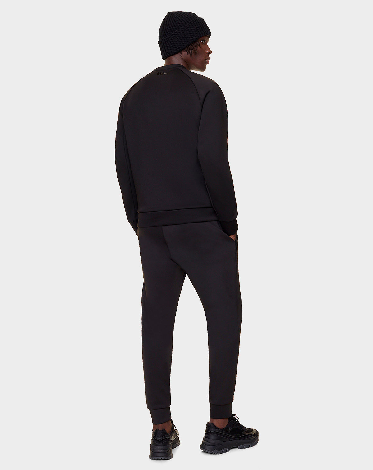 A man wearing the Rudsak DEVAN - 9522510 BLACK MEN crewneck sweater, paired with black jogger pants and a beanie, stands facing away from the camera on a plain light gray background.