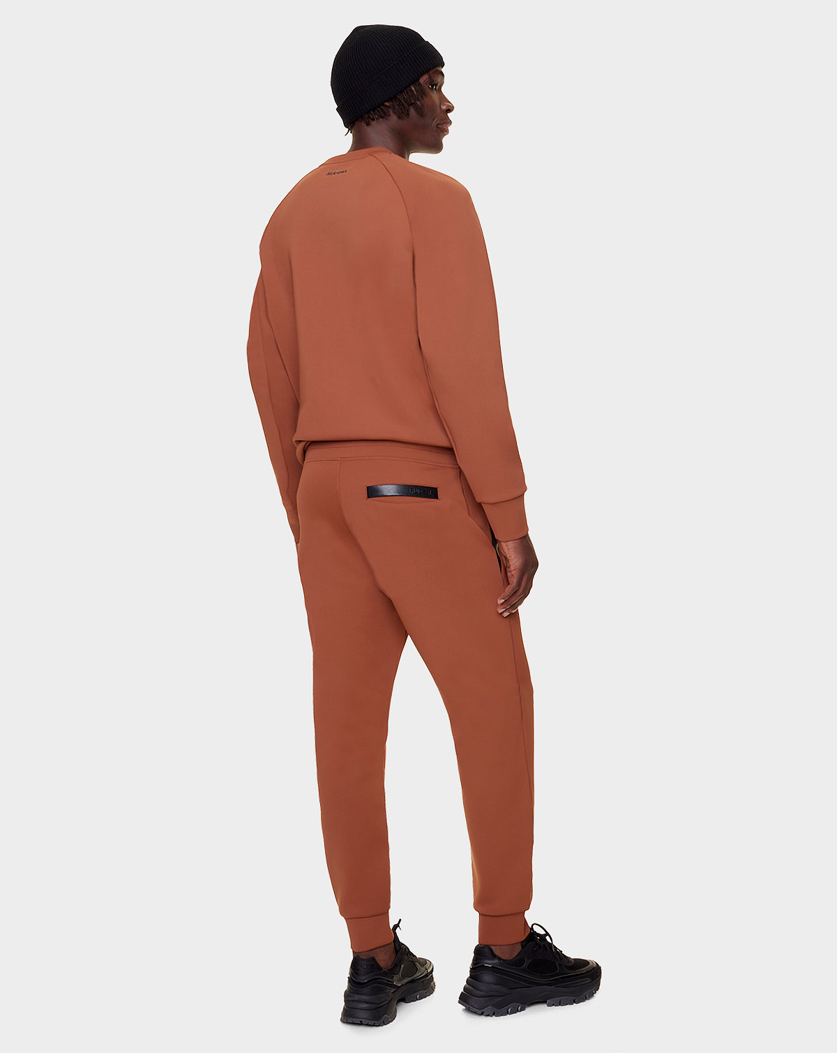 A person wearing the Rudsak DEVAN - 9522510 COHIBA MEN rust sweater tracksuit, black beanie, and black sneakers stands facing away, showcasing the back against a plain light gray background.