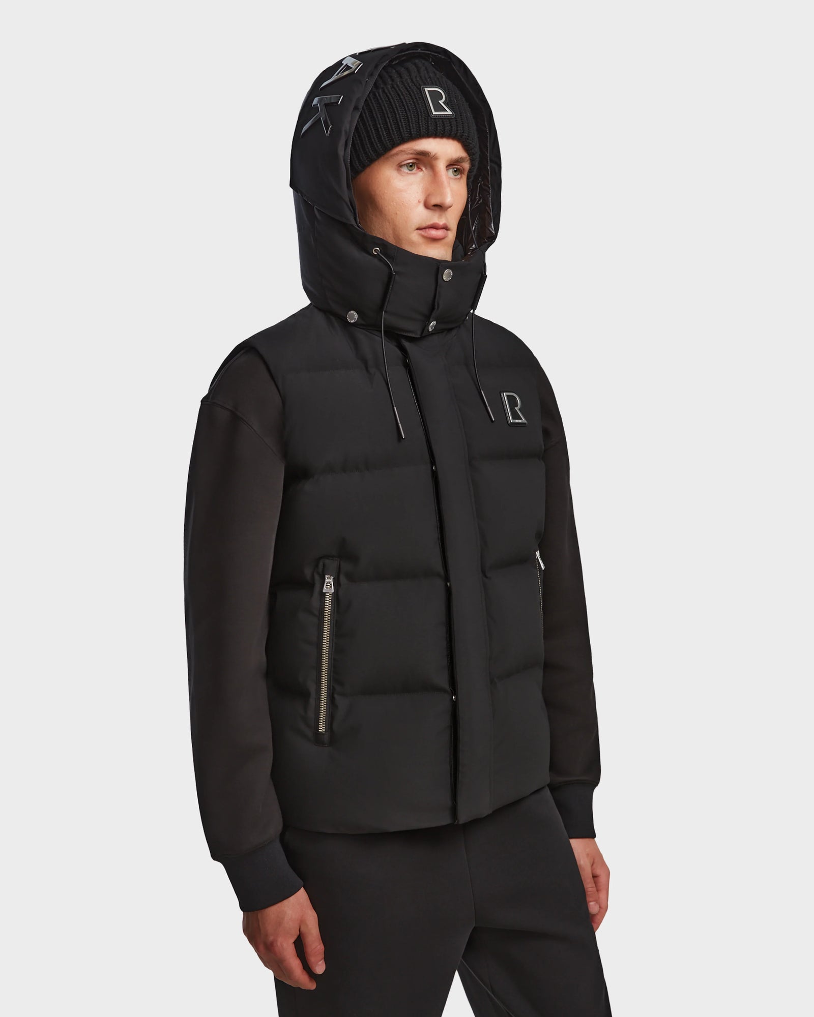 A person wears the Rudsak REN M - 9123512 Black M weather-resistant hooded puffer jacket with zip pockets and a black beanie with white embroidered letters, standing against a plain white background.
