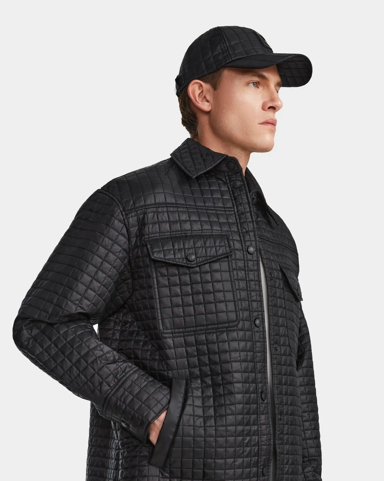 A man in the Rudsak KERRY - 9124003 Black M quilted shirt jacket with eco-friendly insulation and a matching cap stands against a plain light background, looking to the side with one hand in his pocket.